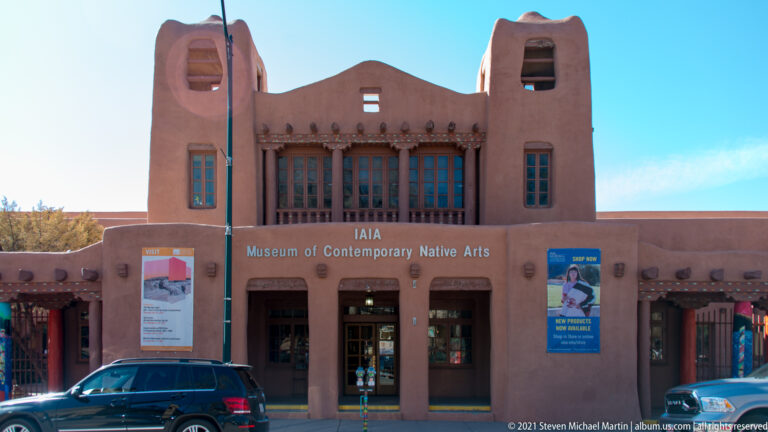 IAIA Museum of Contemporary Native Arts façade with central upper window, entrance bays, posters, and a parked vehicle.