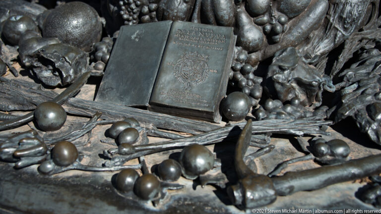 Bronze base detail shows an open book surrounded by rounded objects and elongated forms.