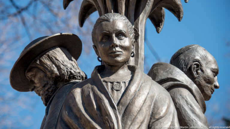 Close view of a bronze group shows a central figure in a collared garment between two others, one wearing a hat.