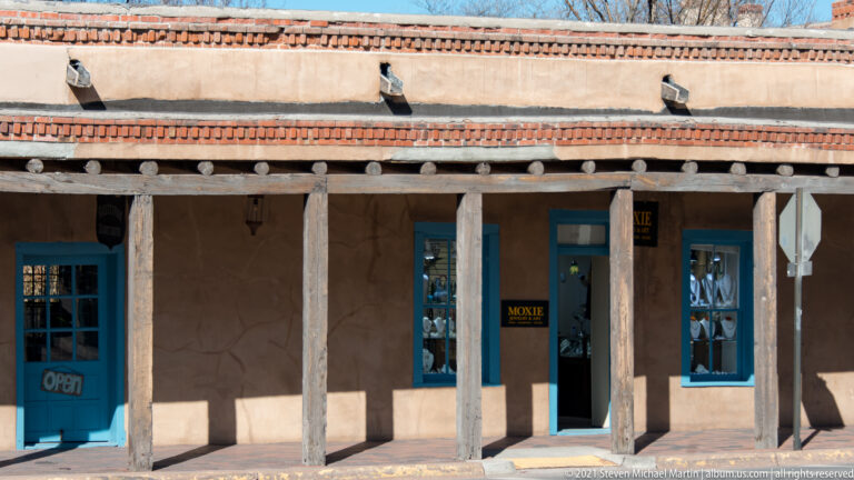 Timber posts support a shaded storefront portal with turquoise-trimmed windows and an open sign.