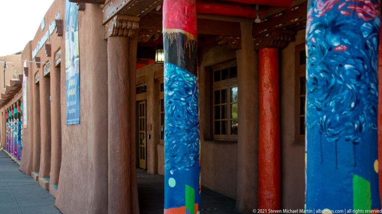 Painted timber columns in blue, red, and green recede along a shaded adobe-style storefront walkway.