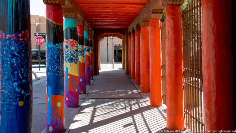 Red-orange posts and a painted ceiling frame a walkway with patterned columns and striped shadows.