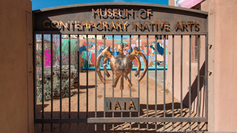 Iron gate reads “Museum of Contemporary Native Arts” above a courtyard sculpture and colorful displays.