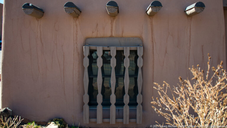 A dark wooden door sits within a shadowed stucco arch beneath four small exterior lights.