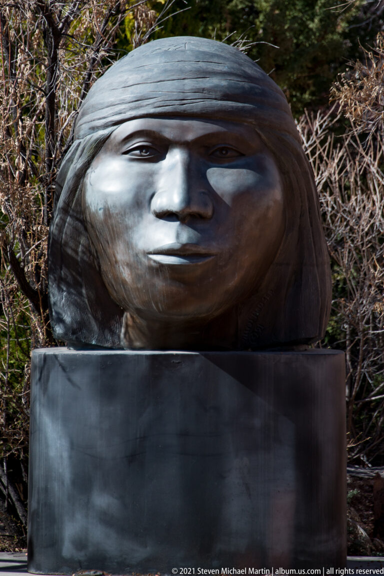 Large bronze head with a broad dark covering sits on a tall pedestal before trees.