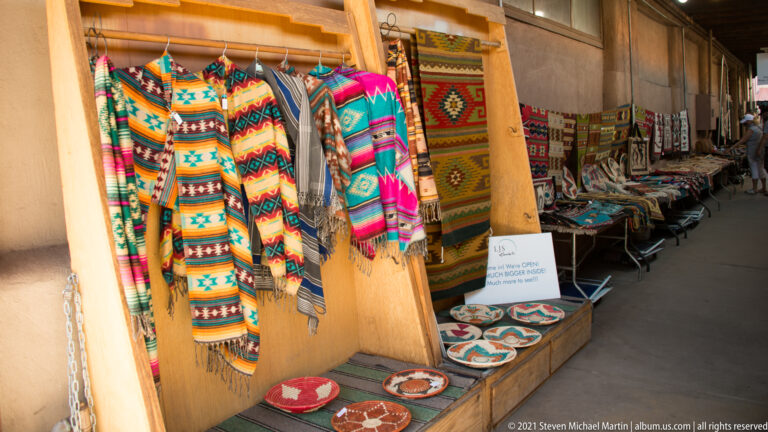 Colorful woven textiles hang above pottery and small crafts arranged along a shaded shop entrance.