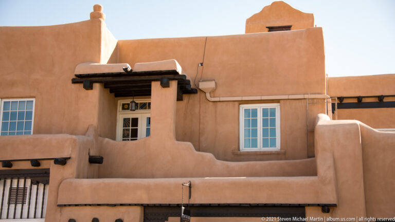 Adobe façade with rounded parapets, projecting wooden vigas, a shaded entrance, and deep-set windows.