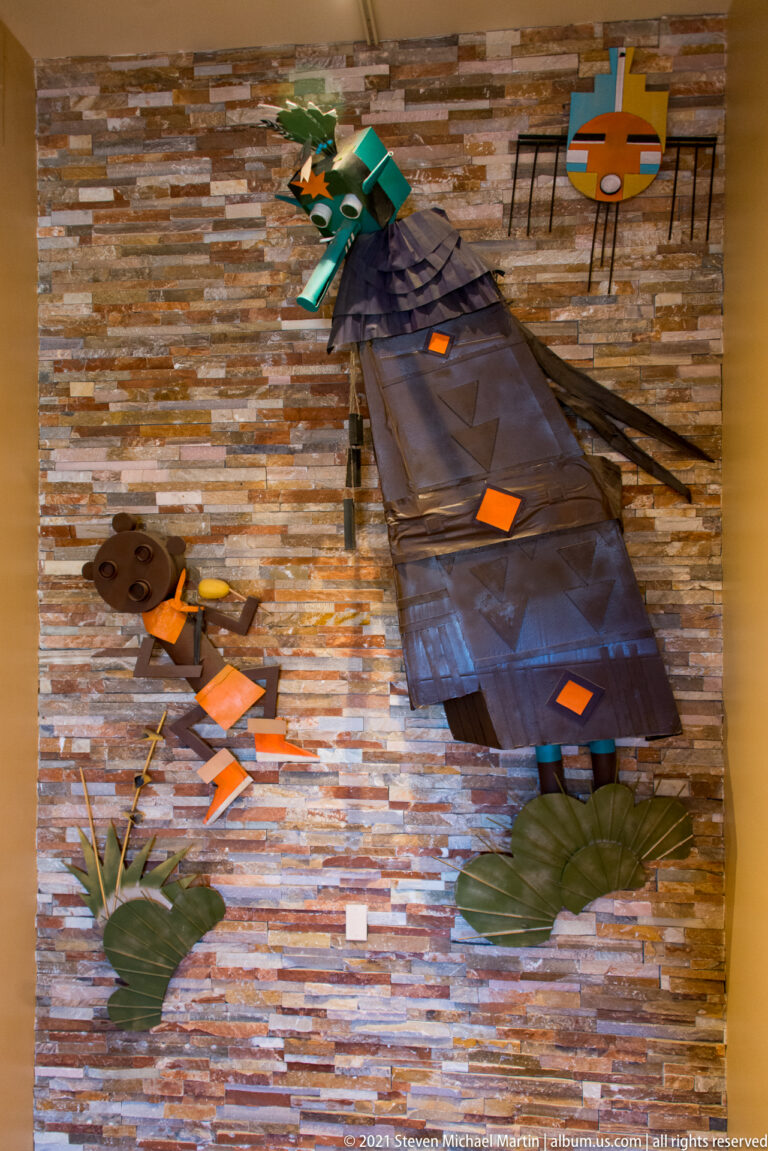 Angular suspended sculpture with a turquoise head and orange accents against a stone wall.