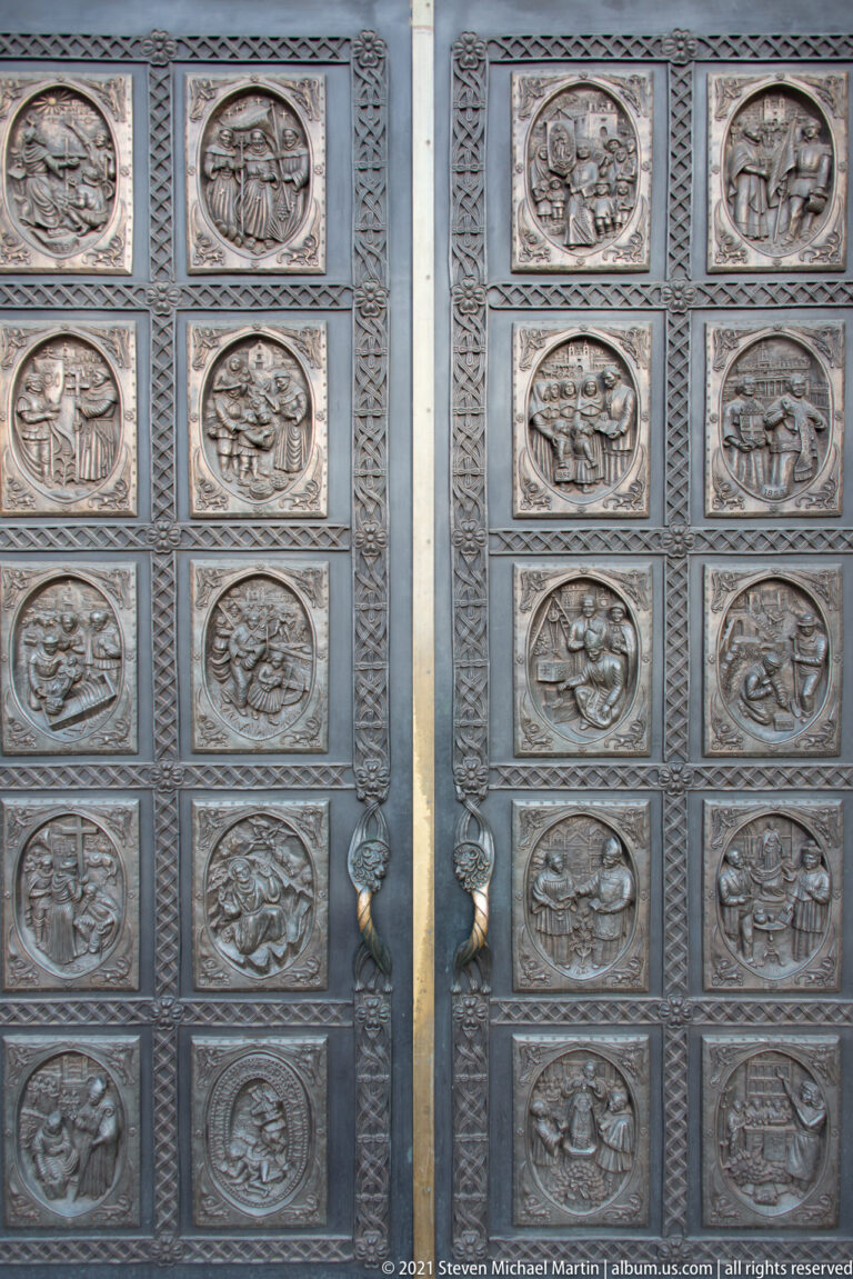 Pair of bronze doors with rows of oval relief scenes, patterned borders, and sculpted central handles.