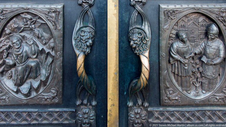 Close-up of robed figures in bronze relief beside elongated decorative handles and curling motifs.