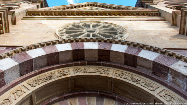 Upward view of a circular stained-glass window above carved stone bands and a pointed church entrance.