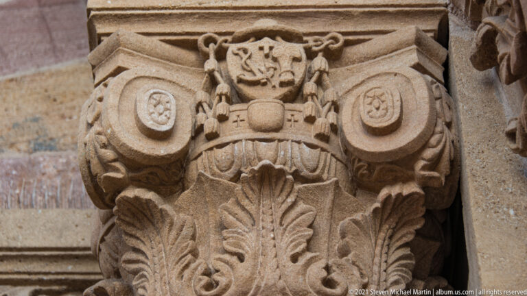 Carved stone capital with curling leaves, paired roundels, and a central shield-like emblem.