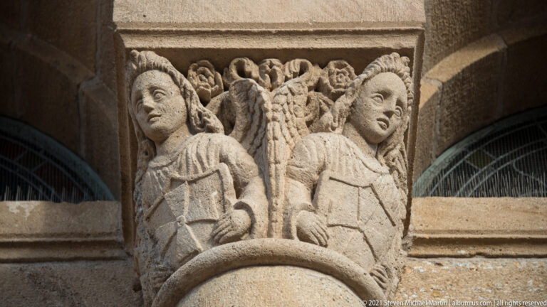 Two seated stone figures face outward from a capital filled with curling foliage.