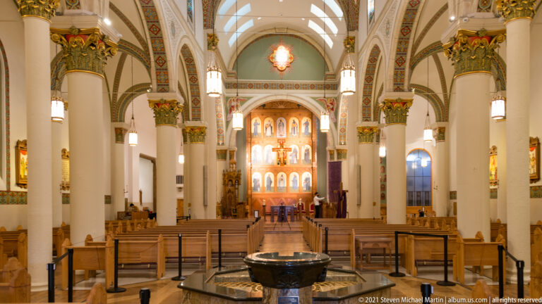 Symmetrical church nave with wooden pews, painted arches, a foreground basin, and a distant sanctuary.