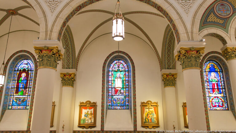 Three stained-glass windows and devotional pictures beneath a painted church vault in the sanctuary.