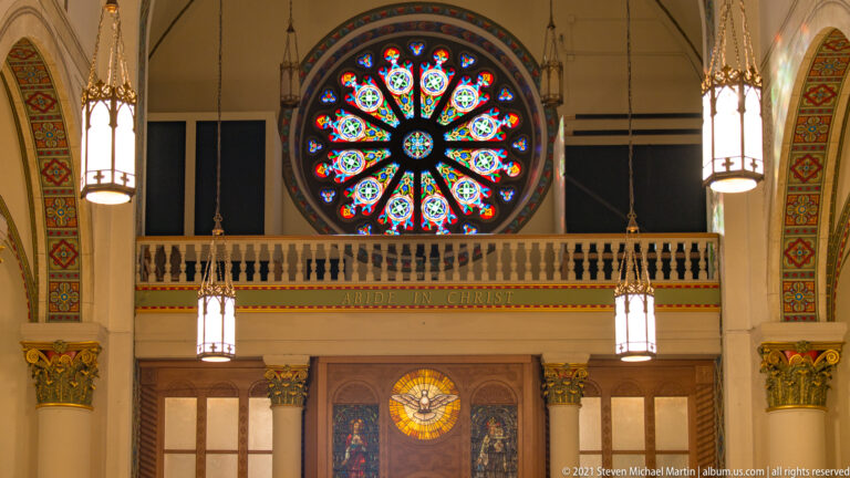 Circular stained-glass rose window centered above a choir gallery between two hanging lanterns.