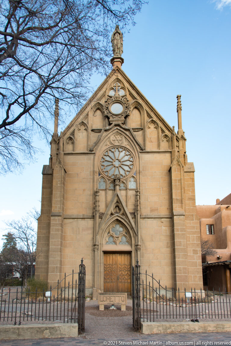 Front façade of a stone church with a pointed entrance, rose window, sculpted gable, and iron fence.
