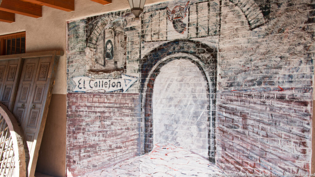 A faded mural surrounds an arched recess, with an arrow pointing toward the words “El Callejon.”