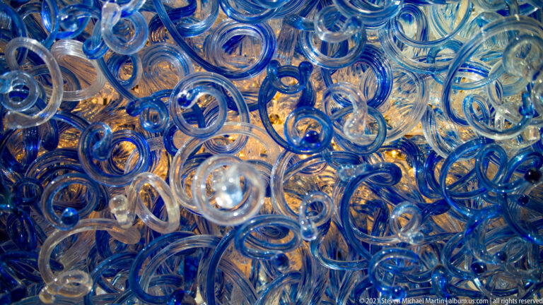 Overlapping loops of transparent and blue glass reflect warm amber light in a dense close-up.