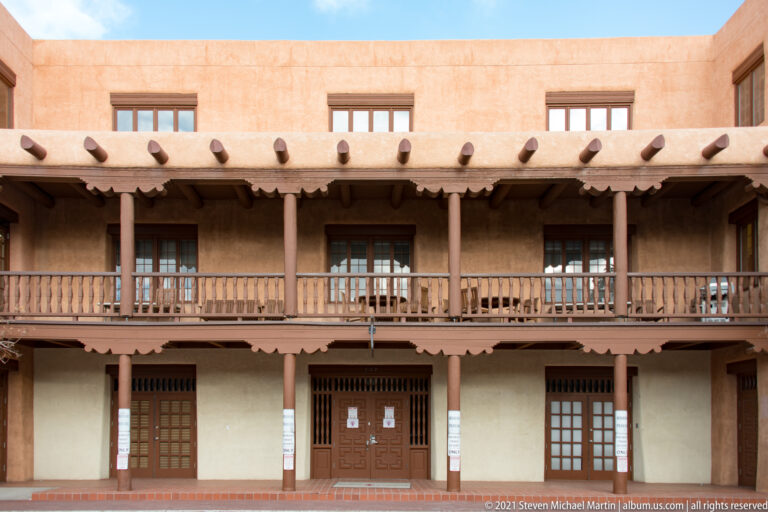 Two-story adobe courtyard façade with a long wooden balcony, evenly spaced posts, and projecting vigas.