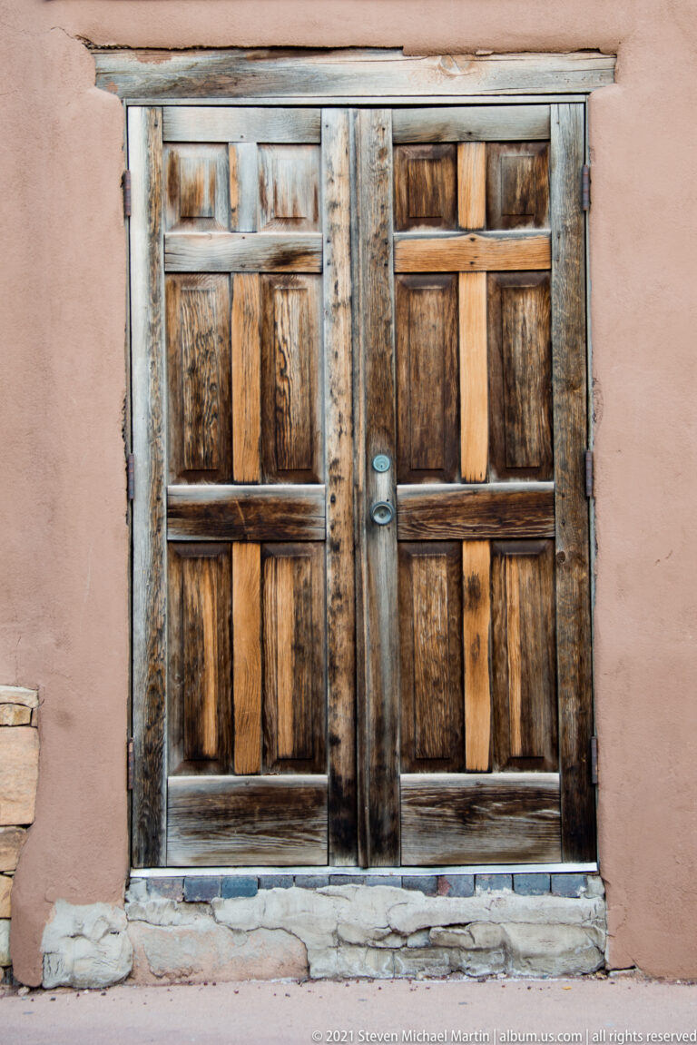 Weathered wooden double doors with horizontal braces and a central latch in a pale adobe wall.