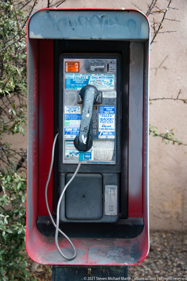 Wall-mounted payphone with a hanging handset inside a red metal hood beside a sidewalk.