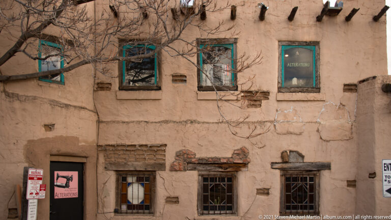 A weathered adobe storefront has repaired openings, turquoise trim, exposed brick, and a small alterations sign.