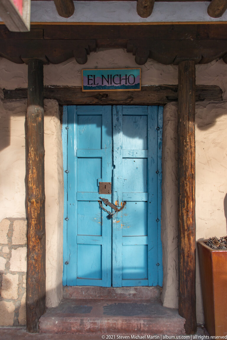 Bright blue double door beneath an El Nicho sign, framed by heavy wooden posts and a shaded porch.