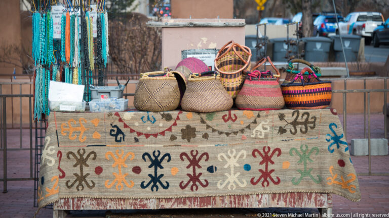 Colorful woven baskets and bags arranged on a patterned table covering at an outdoor street stall.