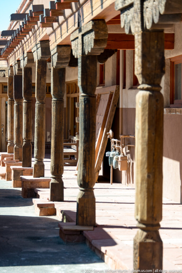 Heavy wooden posts support a shaded sidewalk arcade with storefronts and benches beyond.