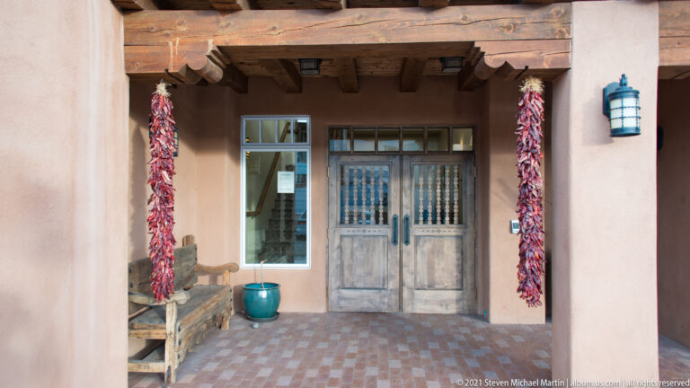 A recessed wooden double door is flanked by red chile ristras, a bench, and a ceramic pot.