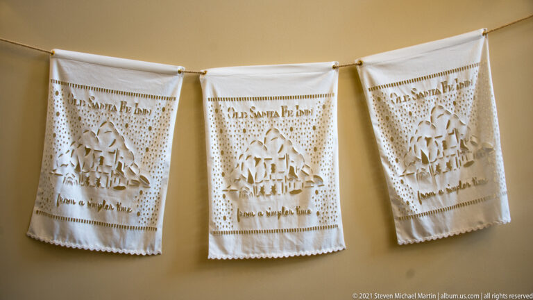 Three cream fabric banners with cutwork designs and lettering hang against a tan wall.