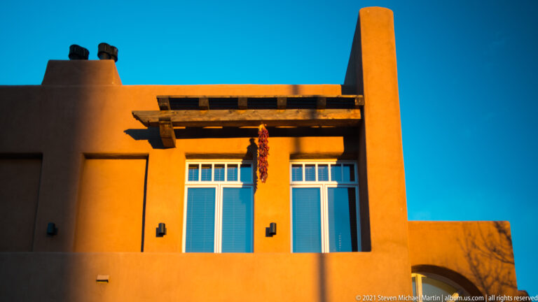 Golden light illuminates orange stucco, blue shutters, timber beams, and a hanging chile ristra.