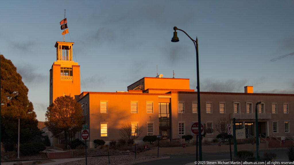 A square tower rises above a low building beside a streetlamp under a cloudy dusk sky.