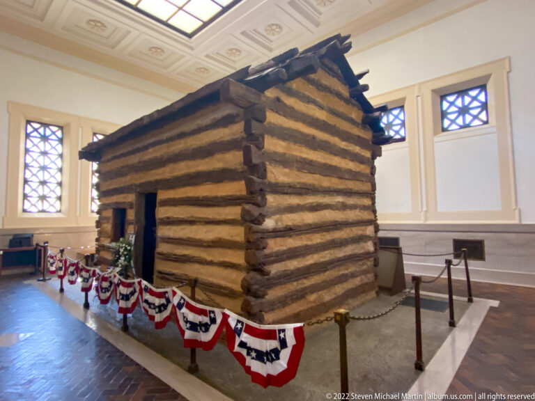 A log cabin stands on a raised platform in a bright rotunda, bordered by patriotic bunting and exhibit panels.