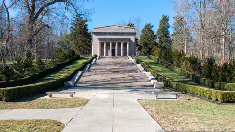 A straight walkway passes paired benches and clipped hedges toward the memorial staircase.