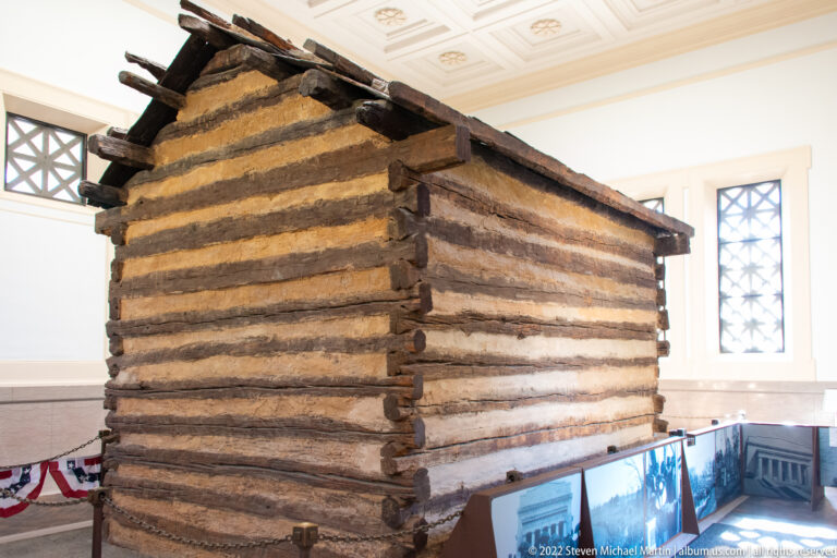 A dark log cabin stands inside a bright memorial hall, with display panels and barriers around its base.
