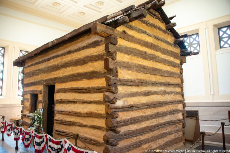 A log cabin with a dark doorway stands behind a rope barrier inside the memorial hall.