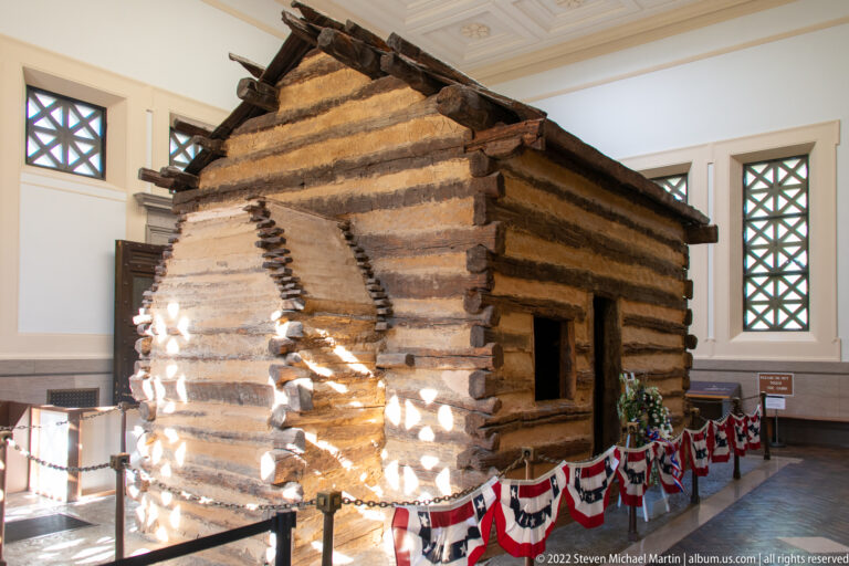 Sunlight crosses a log cabin’s corner and open doorway inside a bright hall with patterned windows.