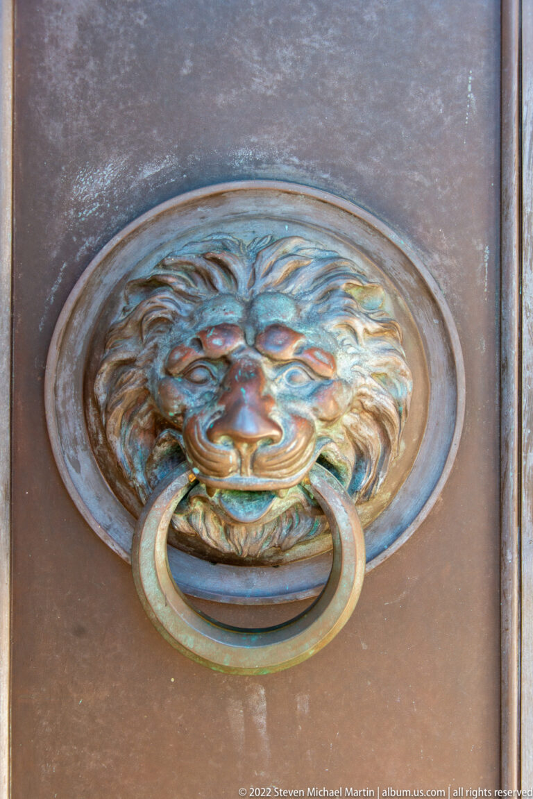 A patinated circular door fitting shows a sculpted face above a large ring handle.
