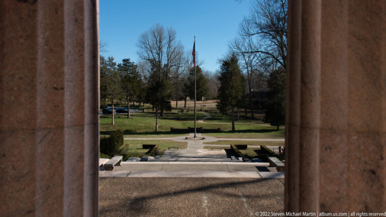 Stone columns frame a flagpole, paths, clipped hedges, and leafless trees across the memorial grounds.