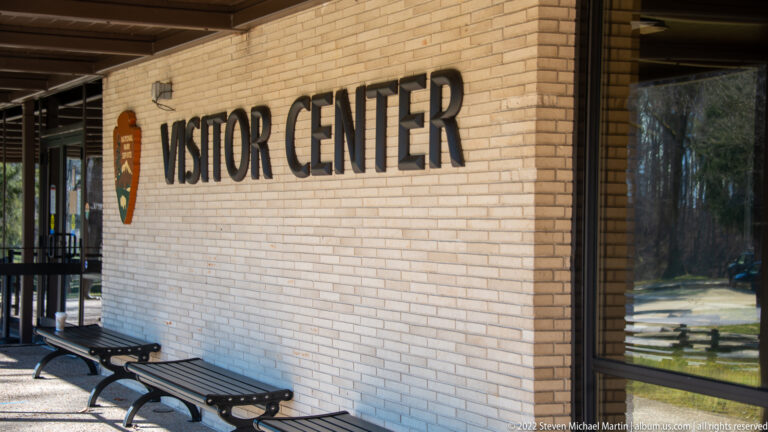 The words “VISITOR CENTER” and an NPS arrowhead are mounted above benches beside glass doors.