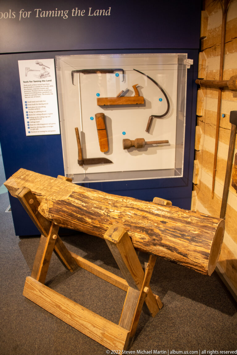 A long wooden implement rests on supports before a case displaying smaller hand tools.