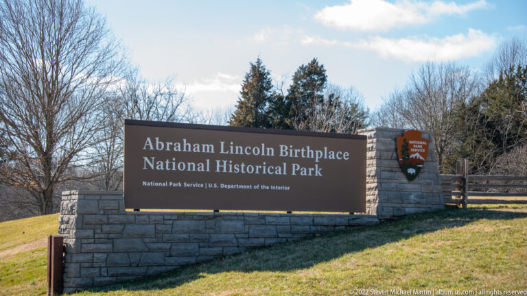 A dark park sign names Abraham Lincoln Birthplace National Historical Park in a stone wall beside a split-rail fence.
