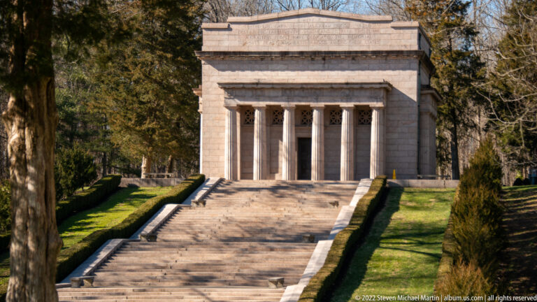 A broad stone stairway climbs to a columned memorial portico framed by clipped hedges and trees.