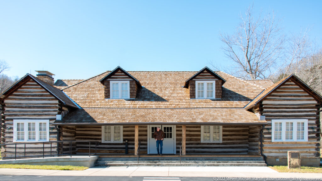 A visitor stands at the central doorway of a broad log cabin with a porch, dormers, and access ramp.