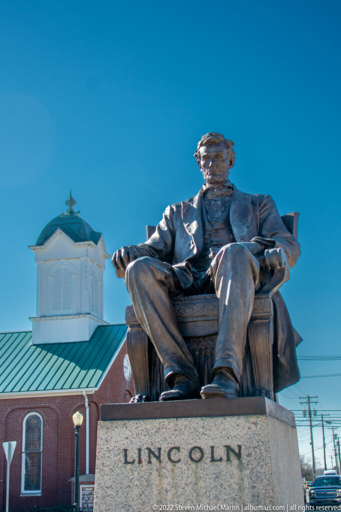 A seated bronze Lincoln statue rises above a stone plinth in front of a brick building and cupola.