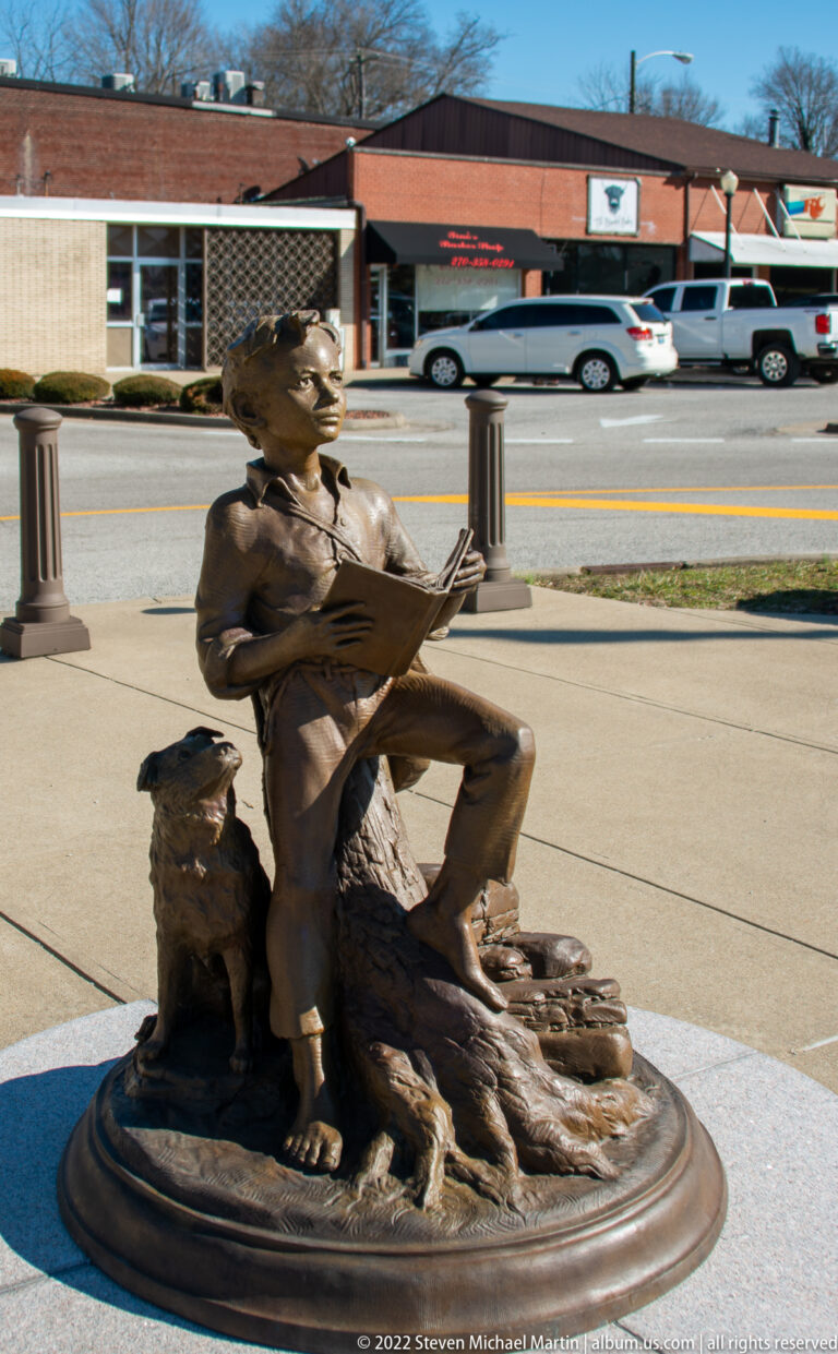 A bronze boy sits on a stump reading an open book, with storefronts and parked cars behind him.