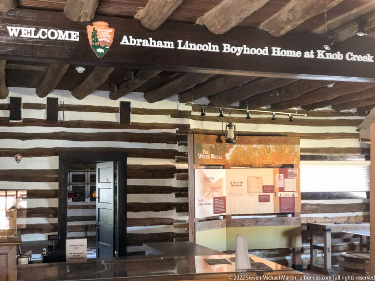 A long exhibit panel hangs on the log wall beneath exposed beams, beside an open doorway and tables.