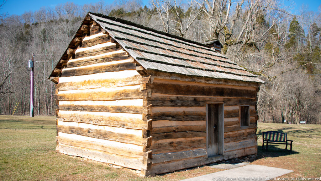 A small log cabin with a steep shingled roof stands on grass before leafless trees and a bench.